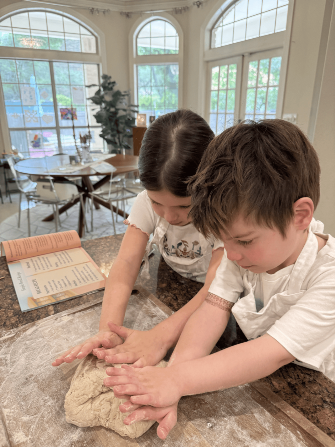 Two children kneading dough together with a Montessori recipe book open nearby